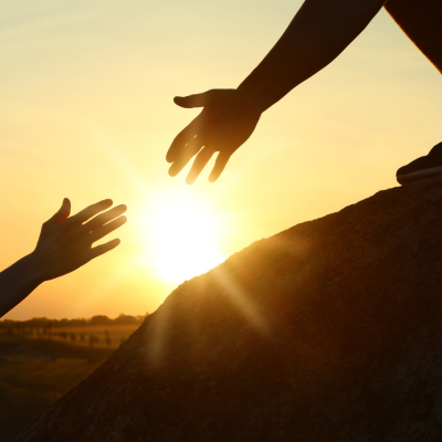 Silhouette of two hands reaching towards each other against a sunset sky, with one hand offering help from above a rock, symbolising support and guidance.